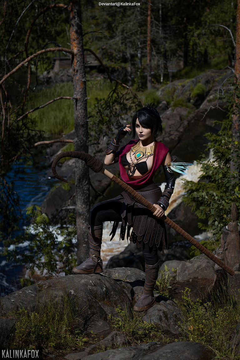 A person in a fantasy-themed costume poses on a rock beside a flowing river, holding a staff, surrounded by trees and greenery.
