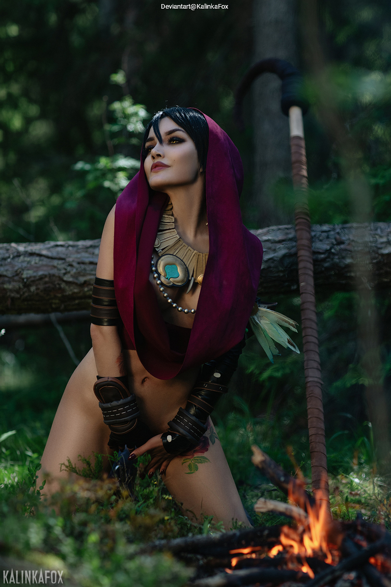A woman in a forest posing in a warrior costume, featuring a purple hood, tribal accessories, and a decorative necklace, with a fire in the foreground.