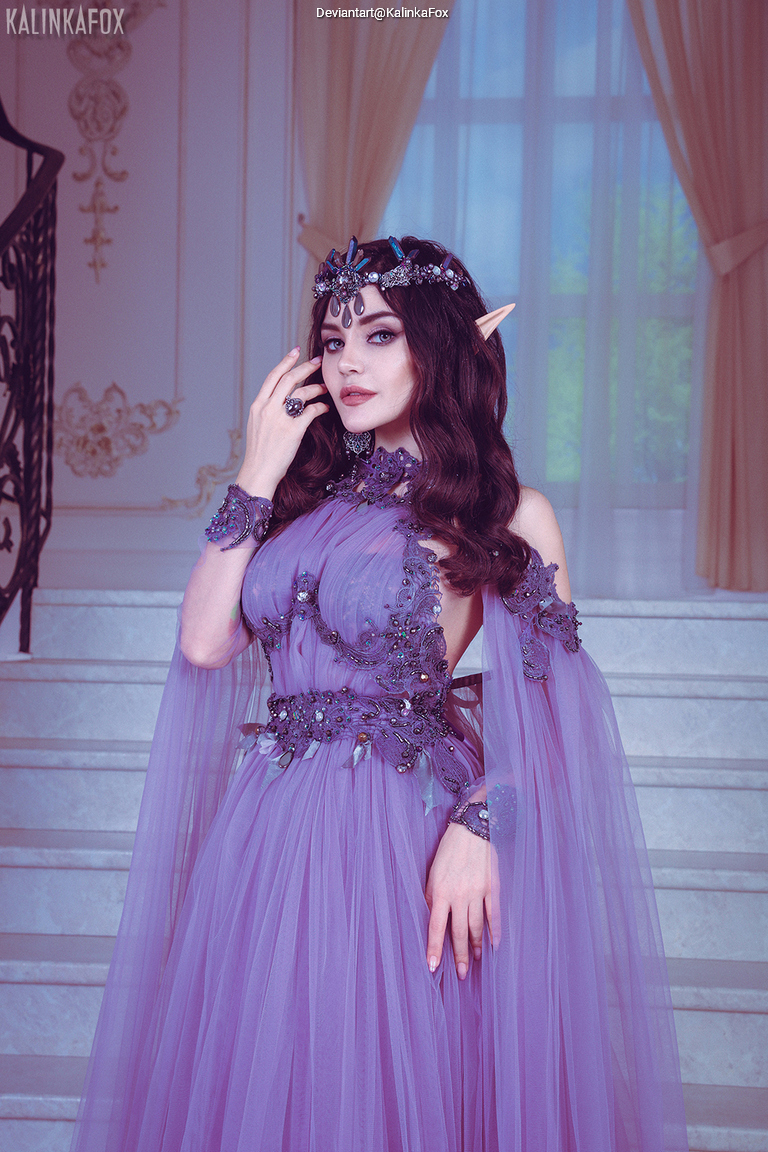 A woman dressed as an elf wearing a stunning purple gown with intricate beadwork, standing gracefully on a staircase with large windows in the background.