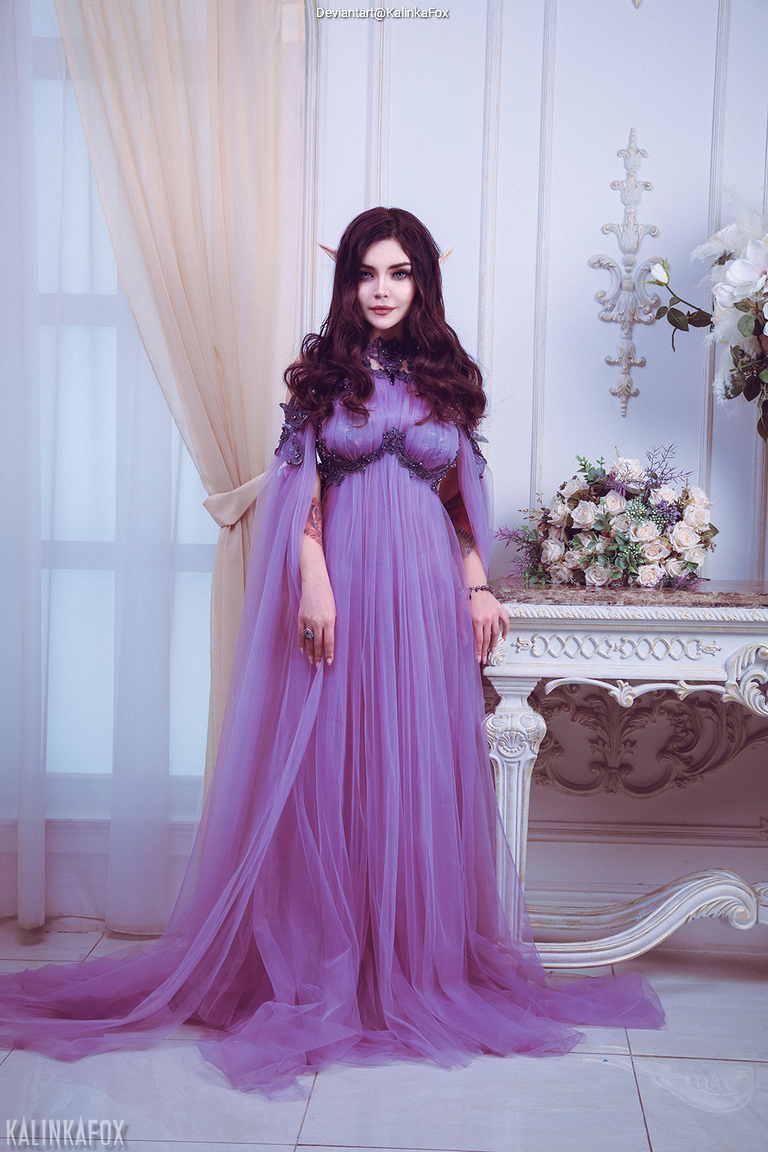 A person with long dark hair and pointed ears, wearing a flowing purple dress, stands elegantly beside a decorative white furniture against a backdrop with soft lighting and floral arrangements.