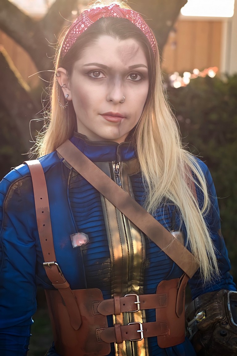 A young woman in a blue and gold costume with a brown chest harness, standing outdoors with a serious expression. She has long blonde hair, a red headband, and subtle makeup with smudged effects.