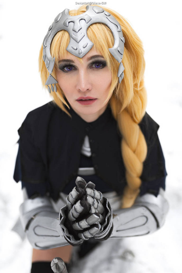 A close-up of a person with long blonde hair styled in a braid, wearing a silver headpiece and metallic arm armor, kneeling in the snow.