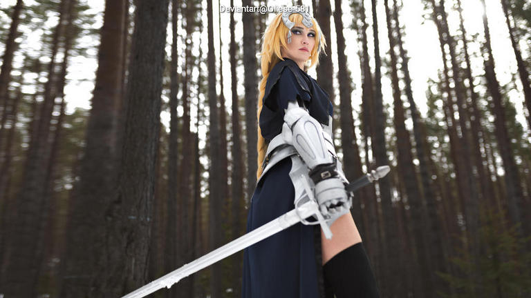 A person in a dramatic pose holding a sword, dressed in a dark outfit with silver armor accents, standing in a forest with tall trees in the background.
