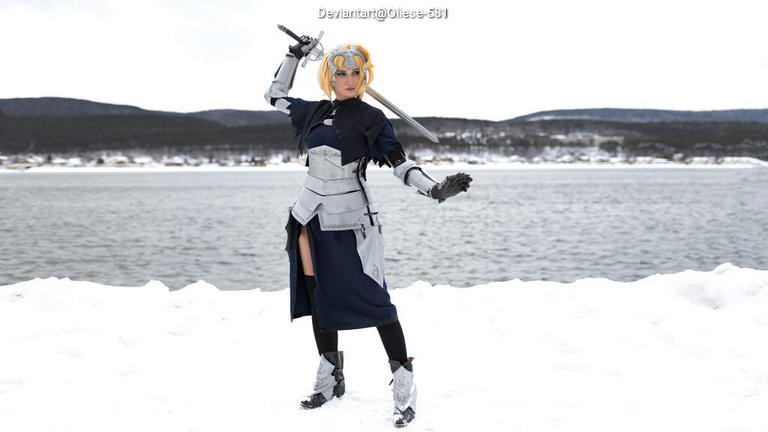 A person dressed in a detailed costume, holding a sword, standing on snowy ground beside a body of water with hills in the background.