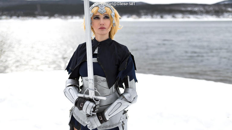 A person in a silver knight costume, holding a sword, standing by a snowy lake with mountains in the background.