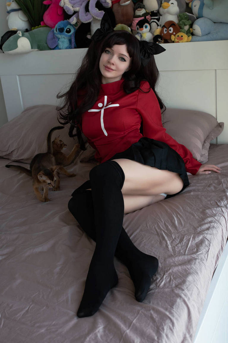 A person sitting on a bed with a playful expression, wearing a red top with a white design and a black skirt. There are plush toys in the background and a cat next to them.