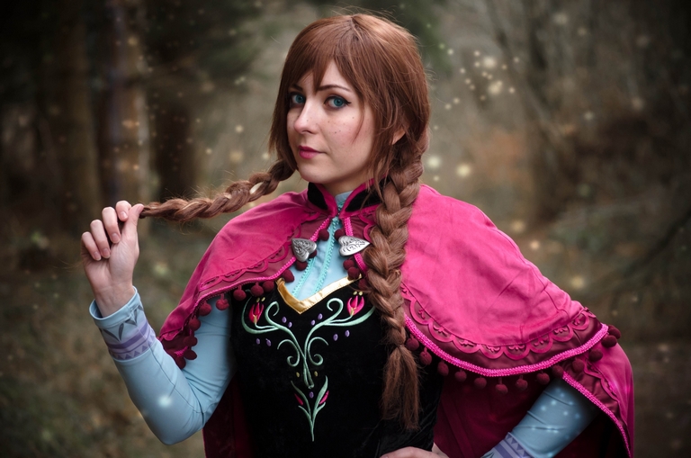 A woman in a vibrant cosplay outfit inspired by a fairytale character, featuring a pink cape and intricate embroidery, stands in a forest setting, playfully tugging on her braided hair.