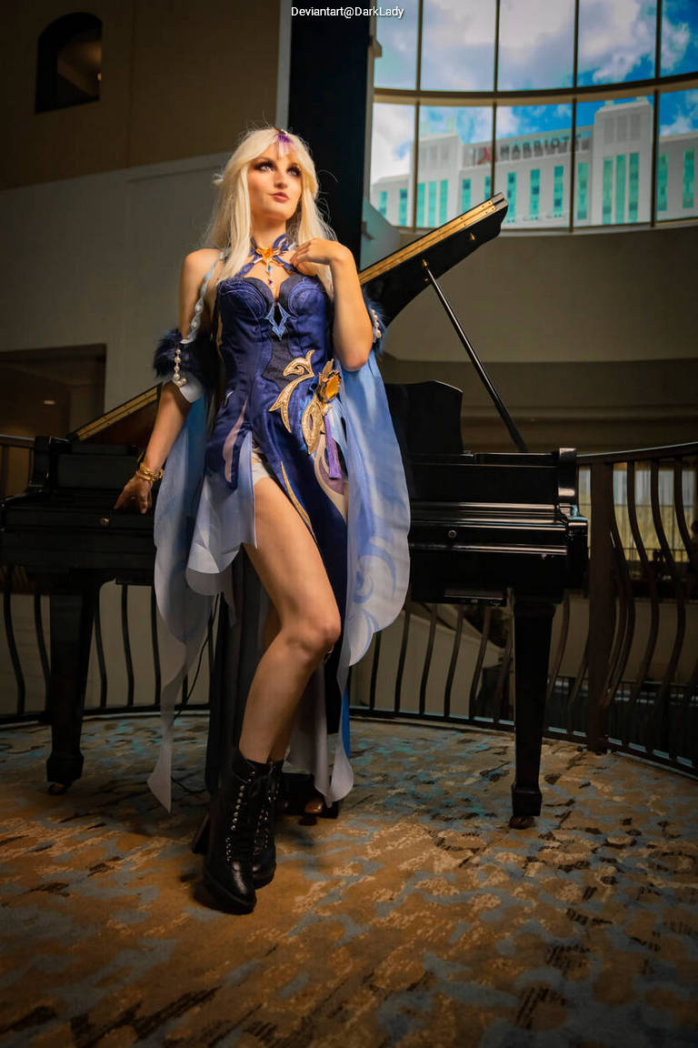 A person in a fantasy costume with long blonde hair, standing next to a grand piano in a well-lit indoor space.