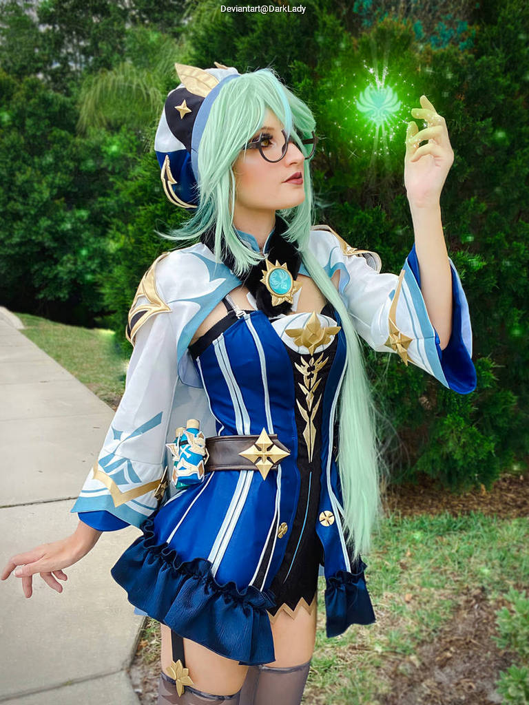 A person in a blue and white fantasy cosplay outfit with long green hair, glasses, and holding a glowing magical object, standing outdoors.