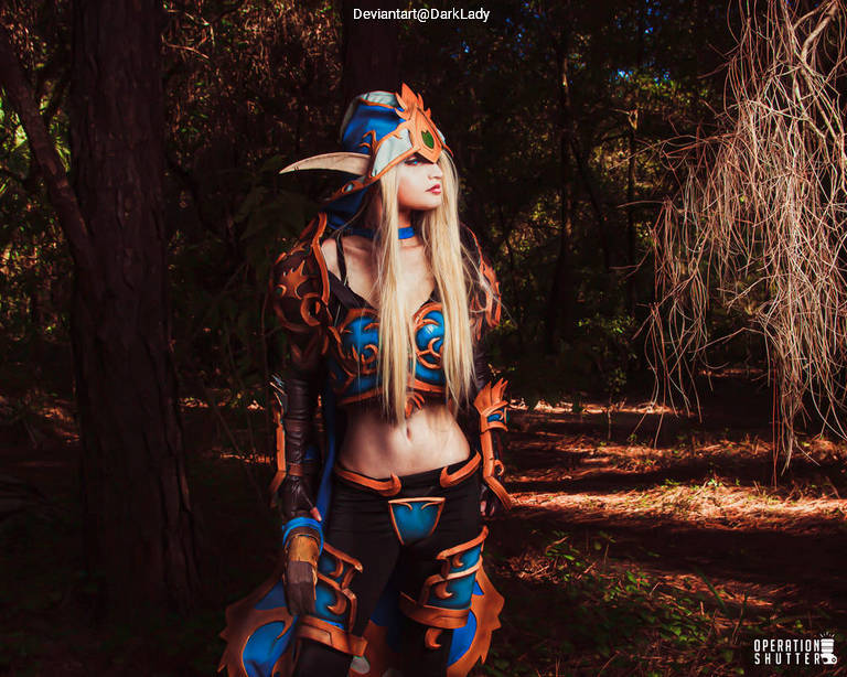 A woman in an elaborate fantasy costume stands in a forest, with blue and orange armor and a unique headpiece, showing long blonde hair.