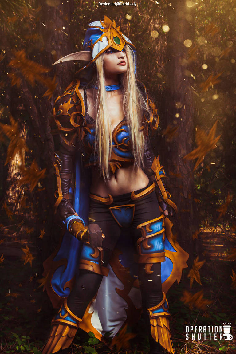 A person dressed in elaborate fantasy armor with blue and gold accents, standing in a forest setting with falling leaves.