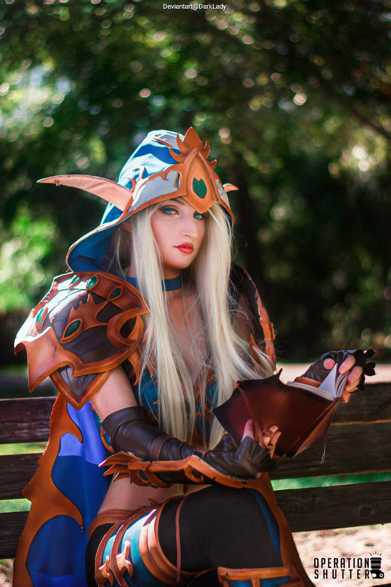A person sitting on a bench dressed in elaborate fantasy cosplay, featuring ornate armor and a blue hood. They are reading a small book, with a serene expression and flowing blonde hair.