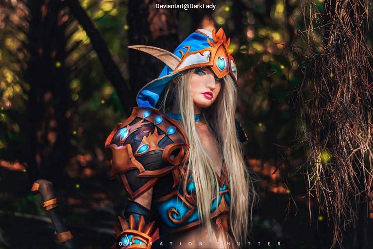 A female cosplayer wearing intricate armor and a large blue hood, posing in a forest setting.