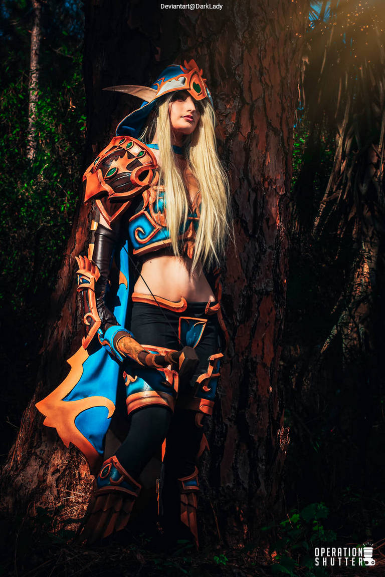 A person wearing a vibrant fantasy-themed cosplay costume, featuring intricate armor and a long wig, stands next to a large tree in a forest setting.