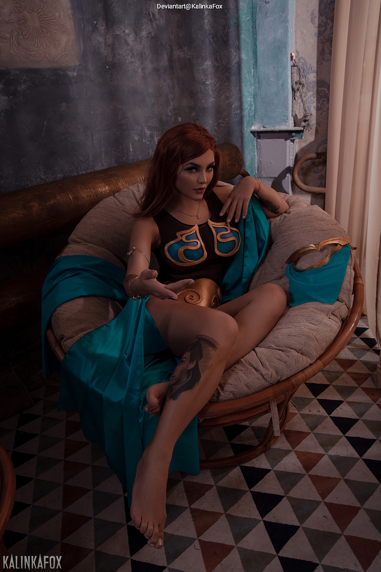 A person with long brown hair is seated elegantly in a large wicker chair, dressed in a stylized costume featuring a blue cape and a top with intricate designs. The background includes textured walls and soft drapery.