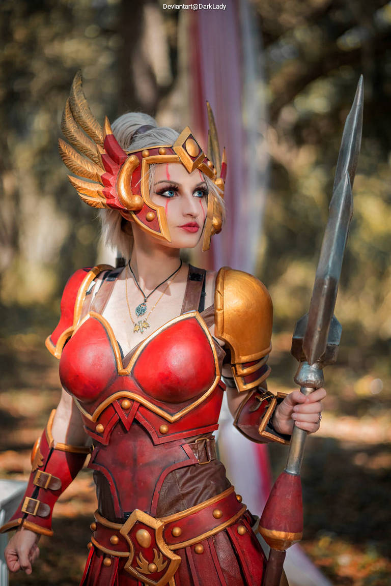 A cosplayer dressed in a detailed warrior costume with golden armor and a feathered helmet, holding a spear, posing in a forest setting.