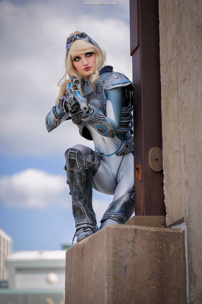 A character in a futuristic costume with silver and blue armor, crouching on a ledge, aiming a handgun while perched against a wall. The background features a cloudy sky and industrial buildings.