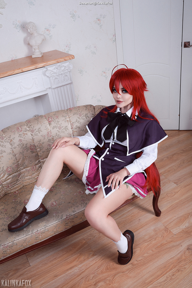 A person with long red hair sitting on a vintage-style sofa, wearing a dark dress with a white shirt and pink pleated skirt, displaying a playful pose.