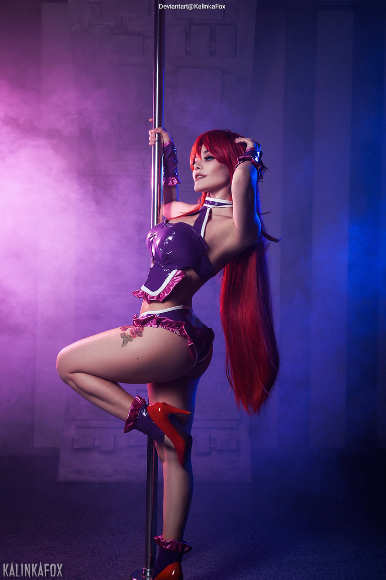 A woman with long red hair poses elegantly while holding onto a pole, wearing a purple costume with ruffled details and red high heels. The background features soft purple and blue lighting with a smoky effect.