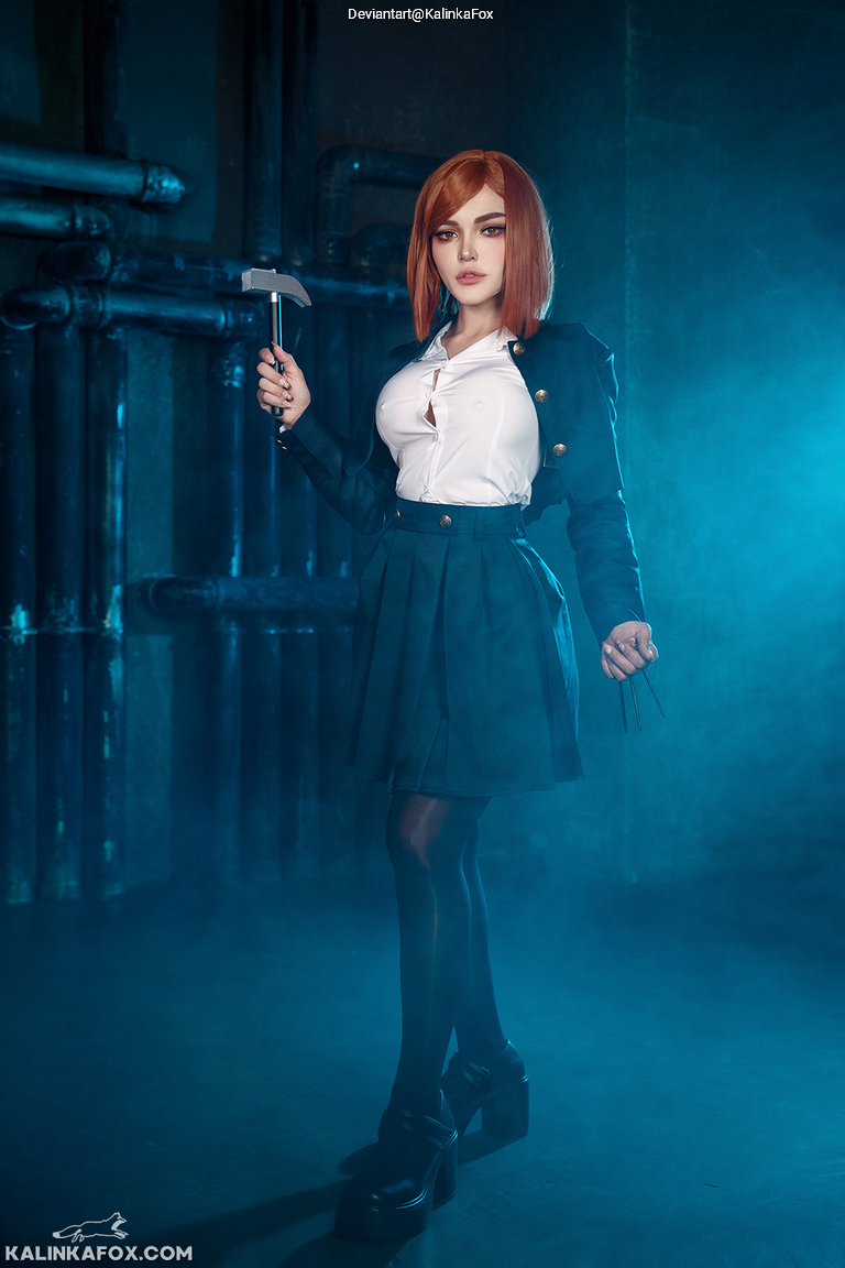 A person with shoulder-length auburn hair, wearing a white blouse and a dark green blazer, stands confidently in a smoky, industrial setting, holding a small hammer.