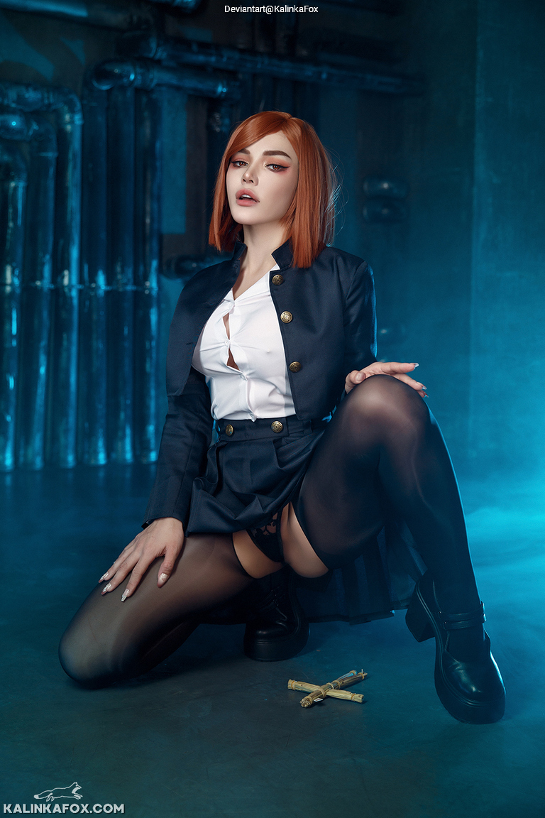 A person with reddish-brown hair wearing a dark jacket and skirt poses against a blue-lit background, sitting on the floor with one leg bent and holding a small object in their hand.