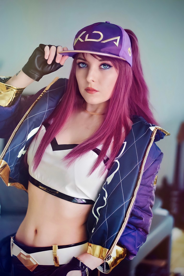 A person with long purple hair wearing a purple cap that says 'KDA', a cropped white top, and a bomber jacket with gold accents. The individual is posing with a hand on the cap, showcasing a confident expression.