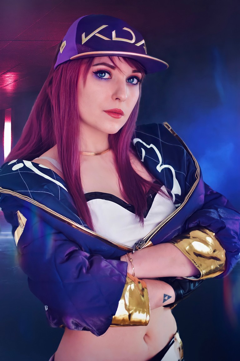 A female cosplayer with long purple hair, wearing a KDA cap and a stylish jacket, poses confidently with her arms crossed in a colorful background.