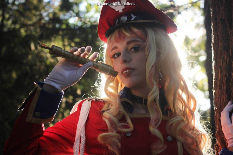 A person in a military-style costume with a red hat and blonde curly hair, holding a baton and posing in a wooded setting.