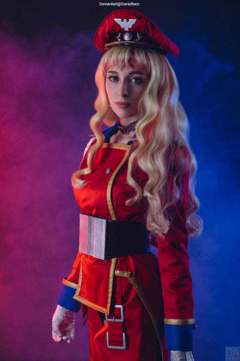 A person in a red military-style costume with a matching hat and long blonde hair, posed in front of a colorful smoke background.