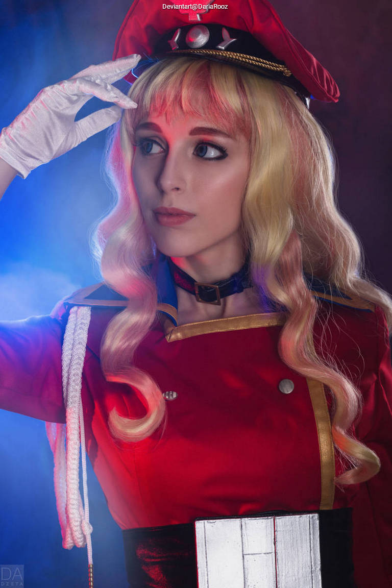 A person in a red military-style uniform and hat, with long blonde hair, posing with a hand raised in a salute. The background is softly lit with blue and purple tones.