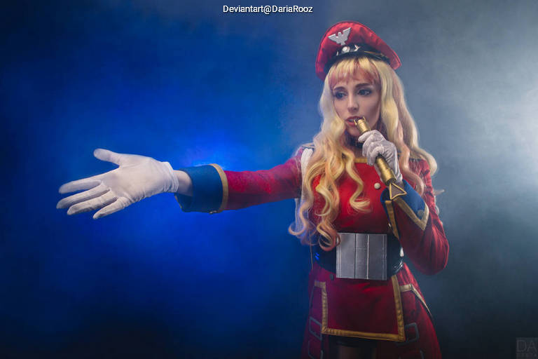 A woman in a military-style red costume and hat poses dramatically, extending her gloved hand and holding a microphone, with a smoky blue background.