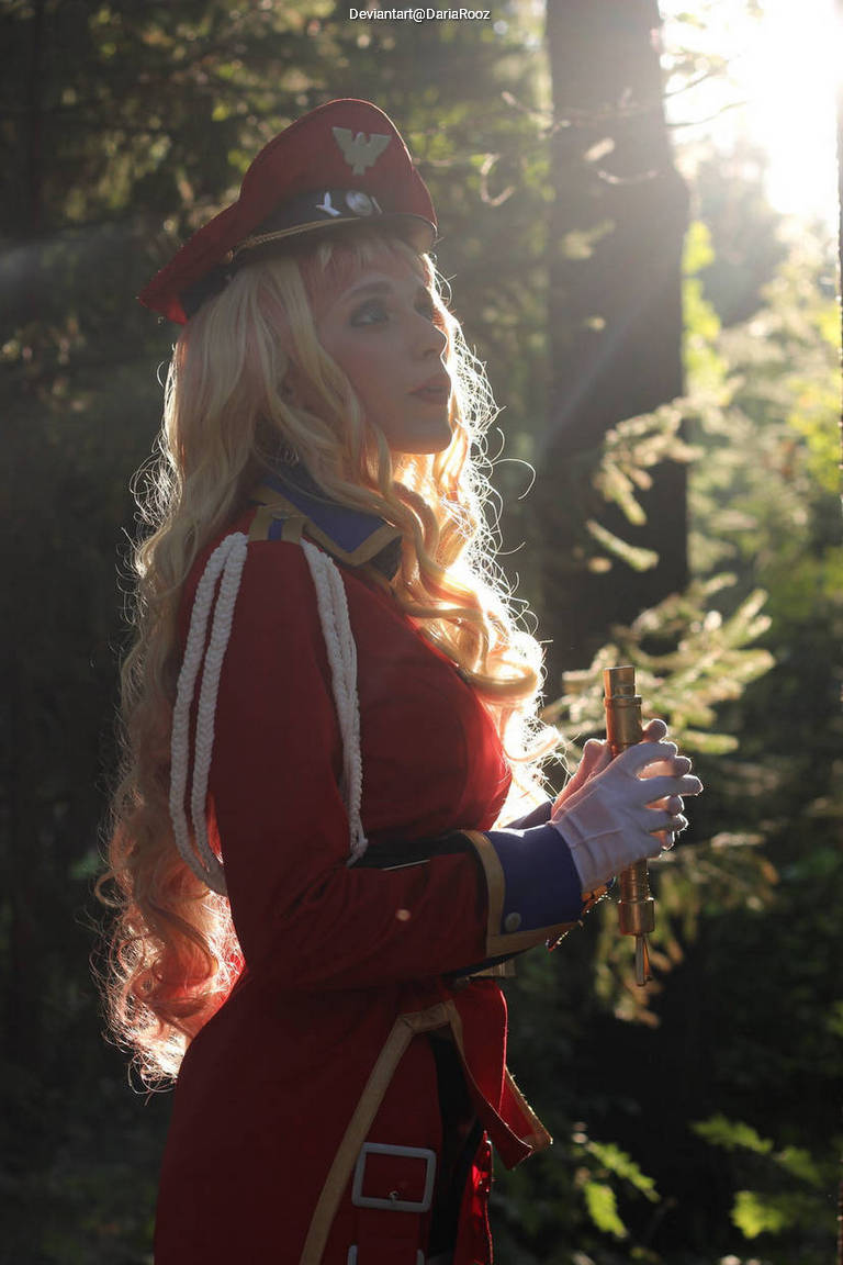 A person in a red military-style costume with a hat and long blonde hair stands in a wooded area, holding a golden object, with sunlight filtering through the trees.