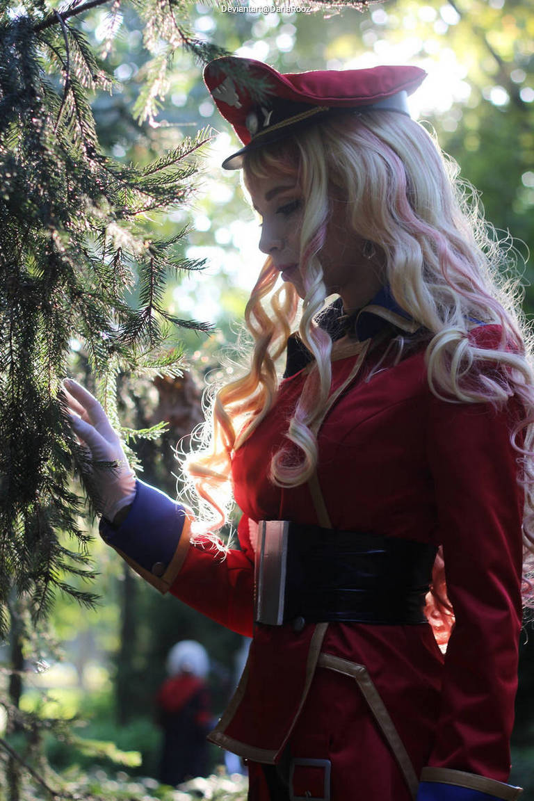 A person in a red military-inspired outfit with a hat, standing in a sunlit forest, gently touching a tree branch.