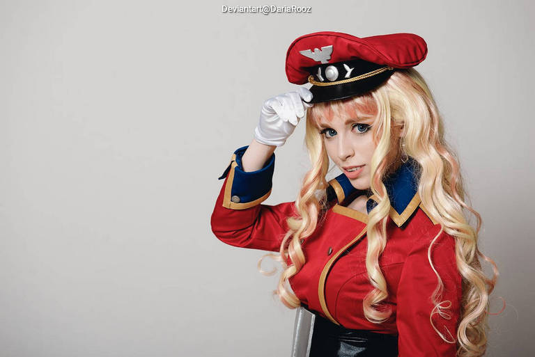 A person in a vibrant red costume, resembling a military or pilot uniform, with long blonde hair and a matching hat. They are posing and adjusting their hat with a smile.