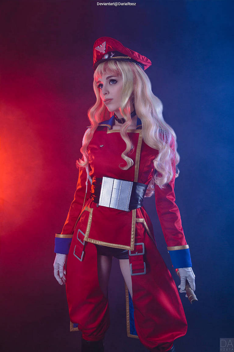 A cosplayer dressed in a red military-style uniform with a matching hat, featuring long blonde hair and posing dramatically against a colorful background.