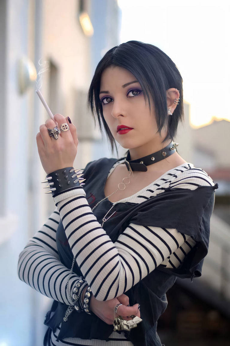 A young woman with short black hair and striking makeup holds a cigarette. She wears a striped long-sleeve shirt layered with a dark vest, accessorized with multiple rings and spiked bracelets. The background features soft, out-of-focus colors that suggest an urban setting.