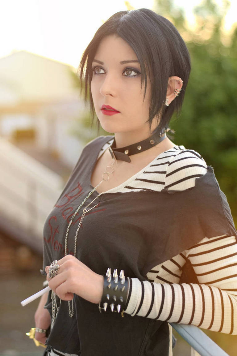 A young woman with short black hair and bold makeup wearing a striped long-sleeve shirt under a black t-shirt, standing outdoors. She accessorizes with a choker necklace and bracelets, holding a cigarette.