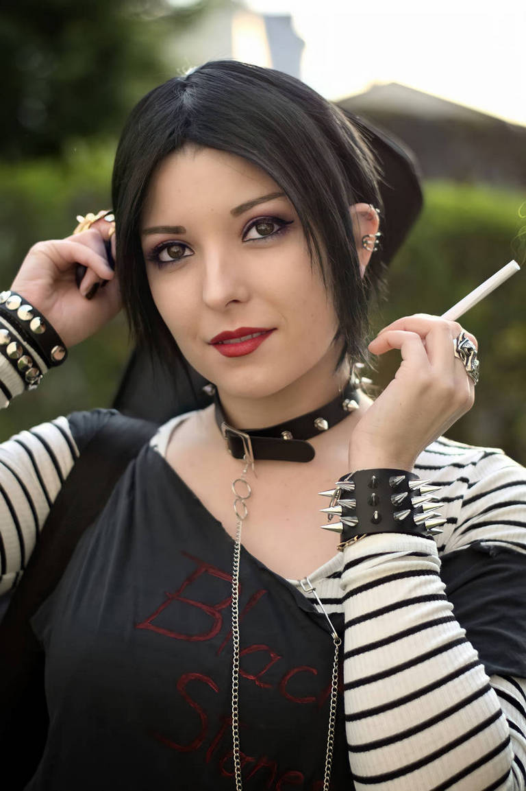 A woman with dark hair and makeup posing with a cigarette, wearing a black shirt and striped sleeves, adorned with studded accessories.