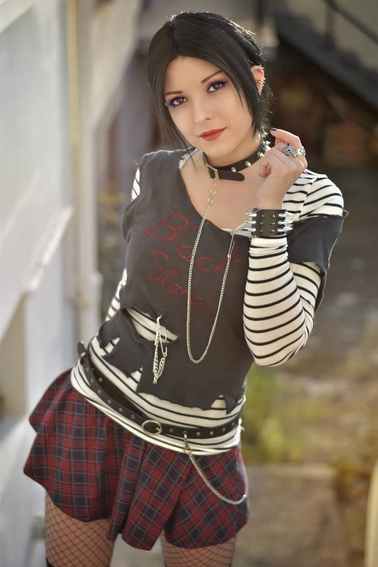 A young woman with black hair styled in a layered cut poses confidently, wearing a graphic t-shirt with red text, a striped long-sleeve shirt underneath, a plaid skirt, and accessories including a choker and bracelets, against an urban background.