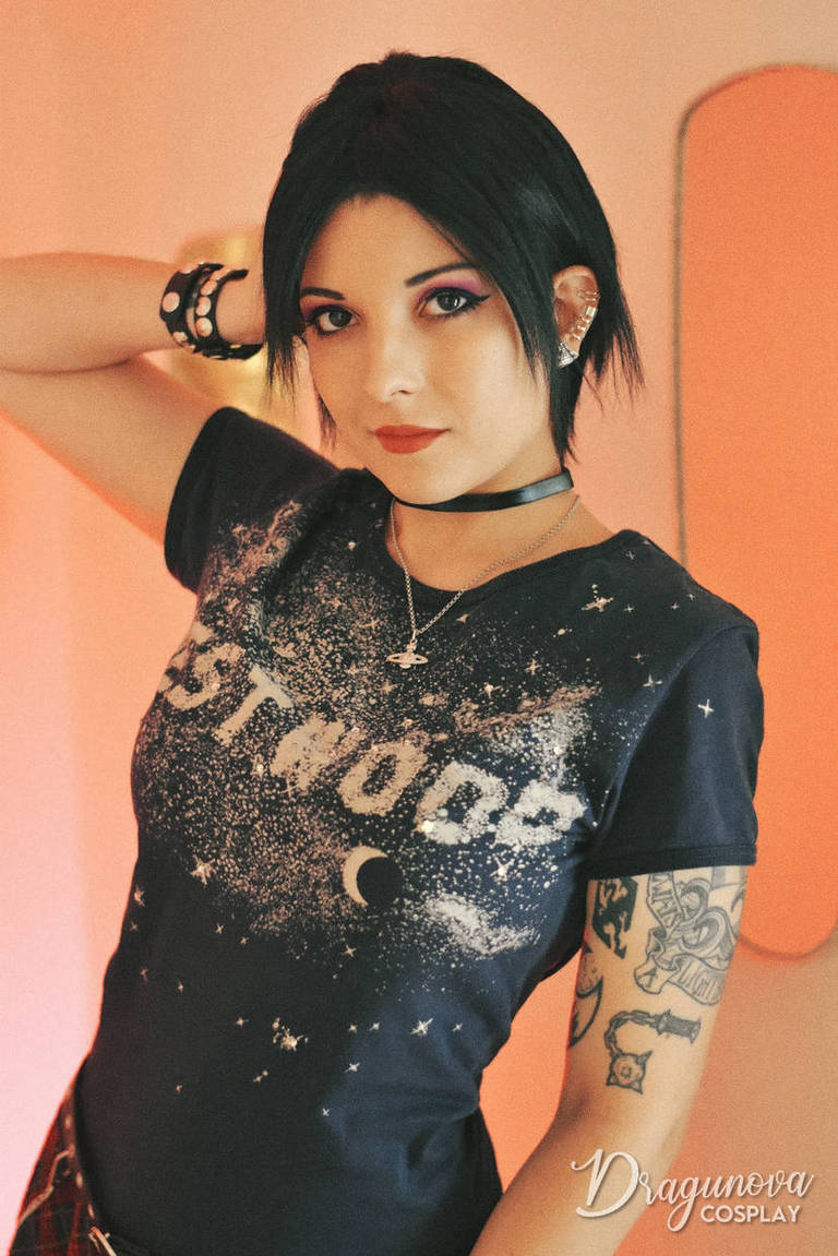 A person with short black hair wearing a navy t-shirt featuring the word 'Westwood' in a starry design, posing with one hand behind their head and a choker necklace.