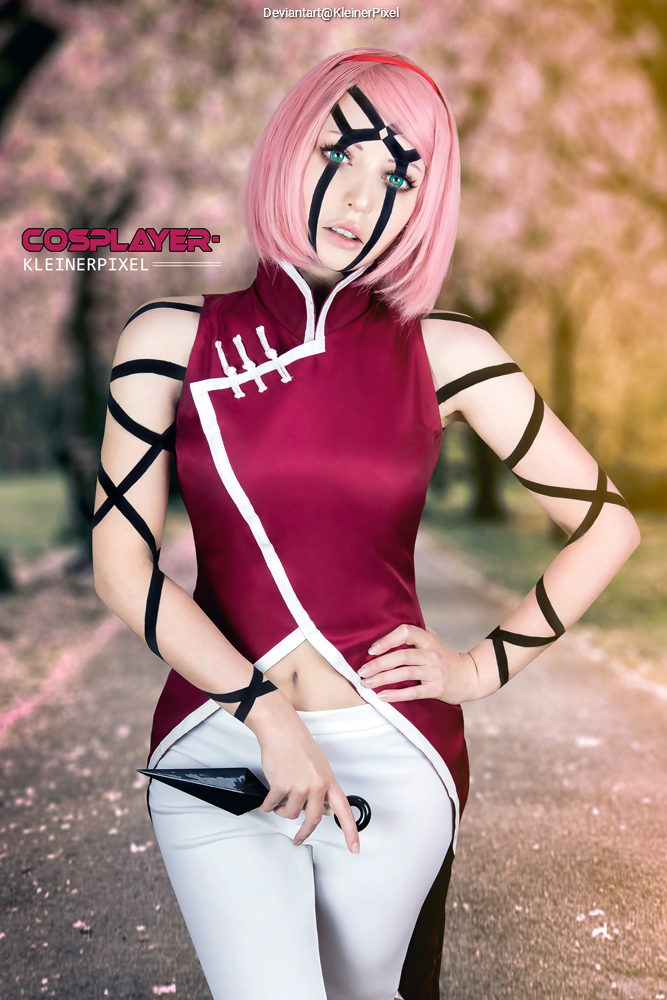 A female cosplayer dressed as a character with pink hair and a red outfit, surrounded by blooming trees in a scenic park.