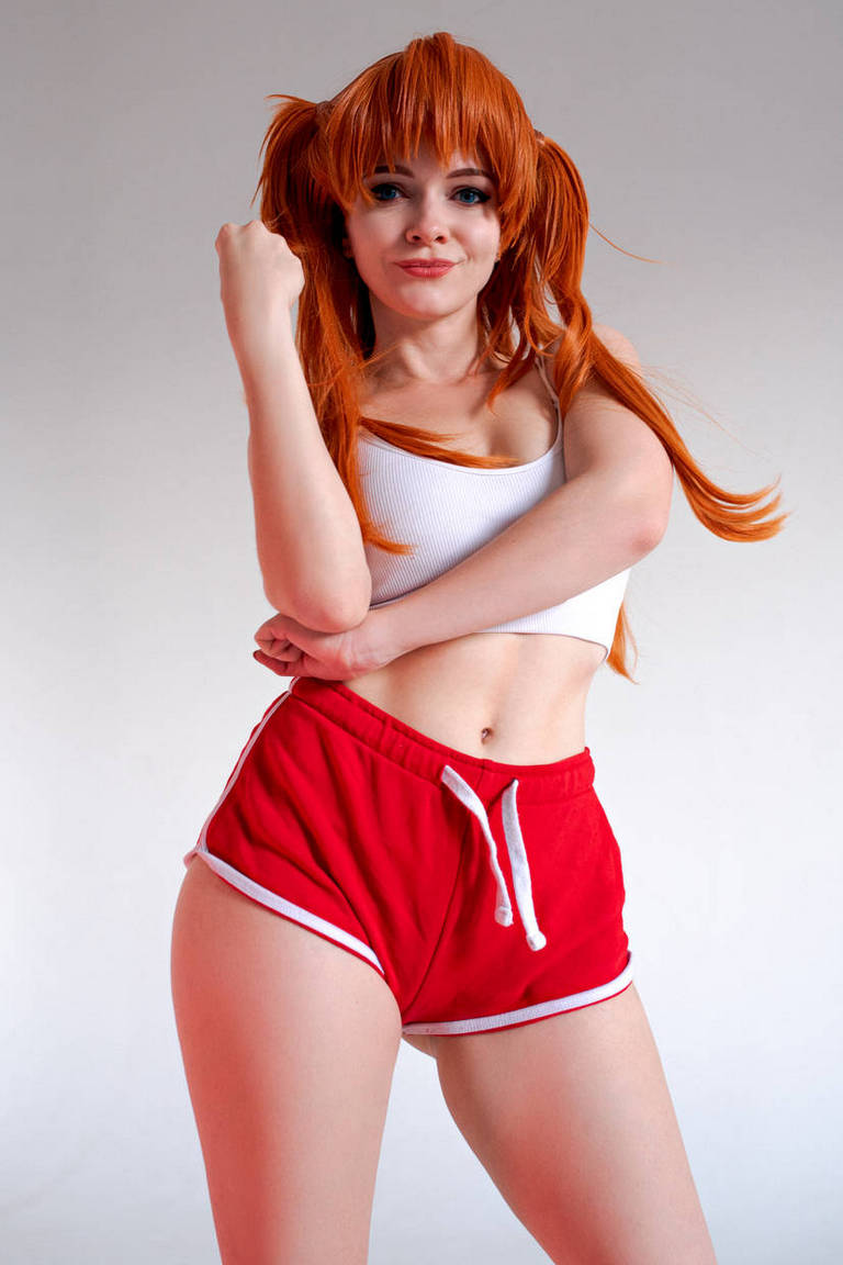 A person with orange hair styled in pigtails is posing confidently in a white tank top and red athletic shorts with white trim.