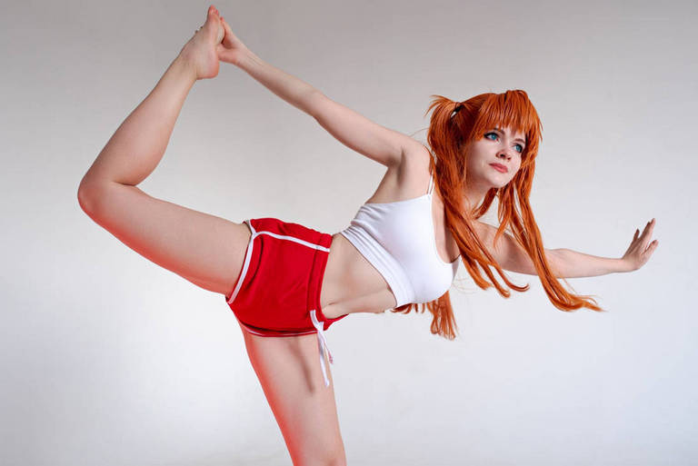 A person with long orange hair performs a dance pose, showcasing flexibility in athletic clothing, including a white top and red shorts, against a plain background.