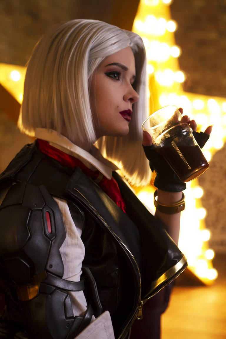 Person in a futuristic outfit with a leather jacket, short white hair, and dark lipstick, holding a glass of beverage, with warm lighting and a star-shaped decoration in the background.