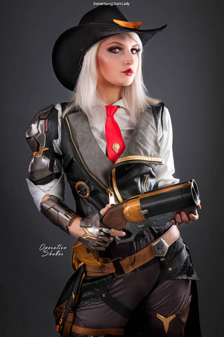 A person dressed in a stylish cowboy outfit with a black hat, silver and gold attire, and holding a large revolver. The makeup features bold red lips and dramatic eye makeup, creating a striking appearance.