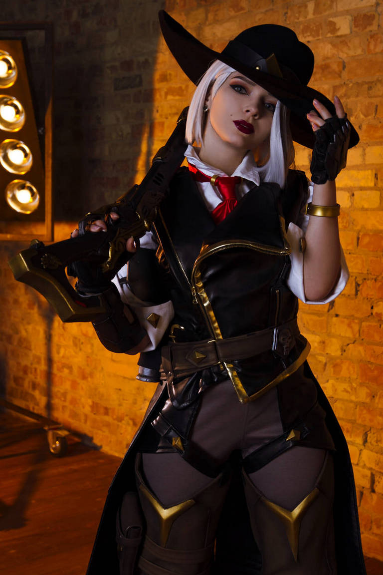 A person dressed in a detailed costume with a wide-brimmed hat, holding a stylized gun. The outfit features a mix of dark and light colors, with gold accents and a red tie, against a brick wall backdrop.