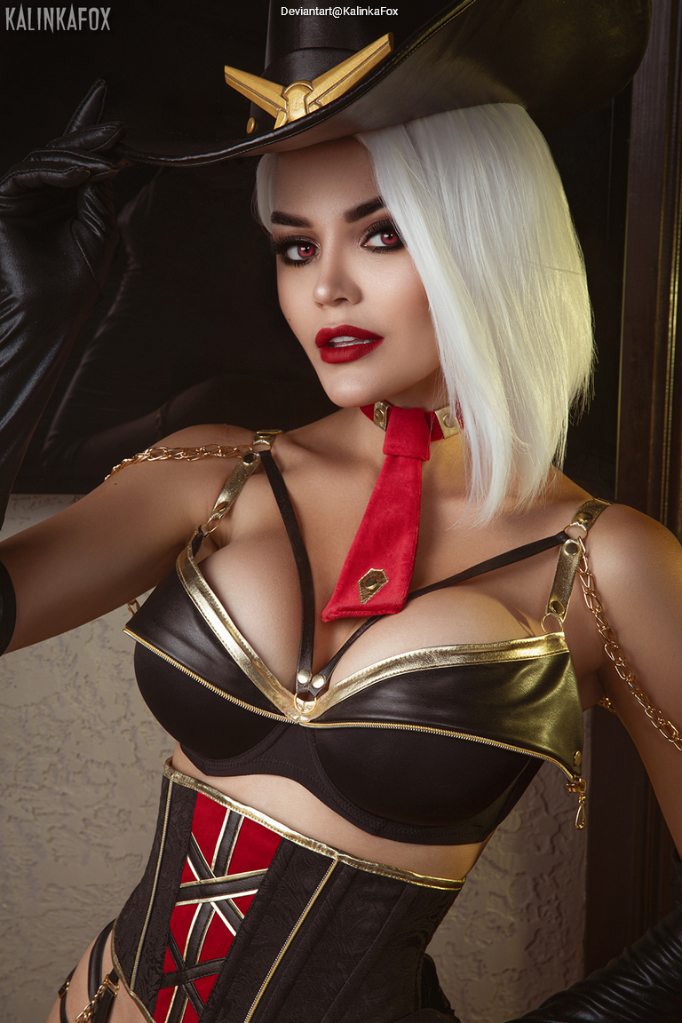 A woman with short white hair poses confidently in a black hat and stylish outfit, featuring a leather bodice and red accents. Her makeup is bold, with dark red lips and defined eye makeup.