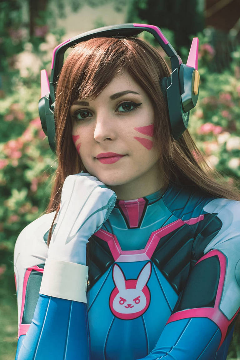 A person in a colorful cosplay outfit resembling a character from a video game, with brown hair and pink accents on their cheeks, posing thoughtfully outdoors.