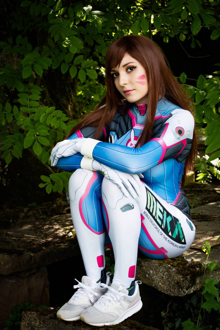 A person in a colorful D.Va cosplay outfit from a video game, sitting on a rock surrounded by green foliage.