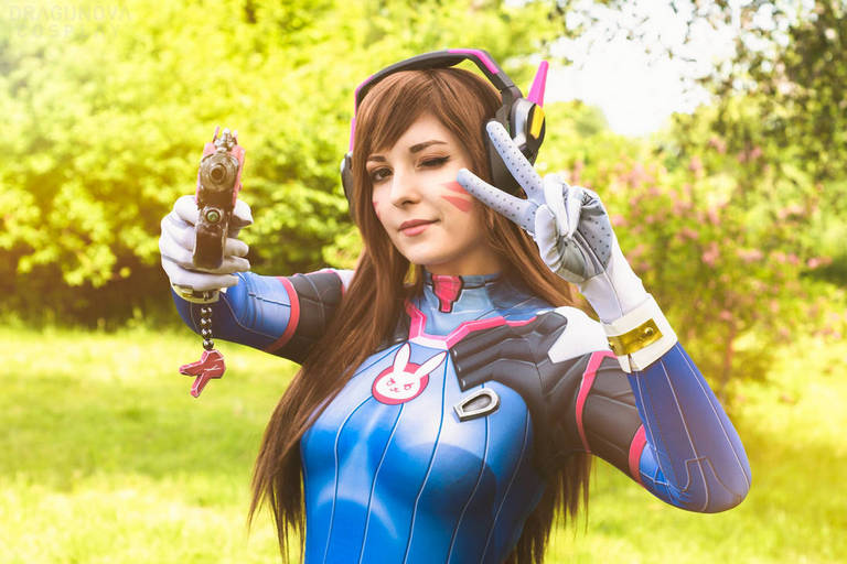 A person in a blue and pink cosplay outfit poses with a toy gun, making a peace sign with one hand in a lush outdoor setting.